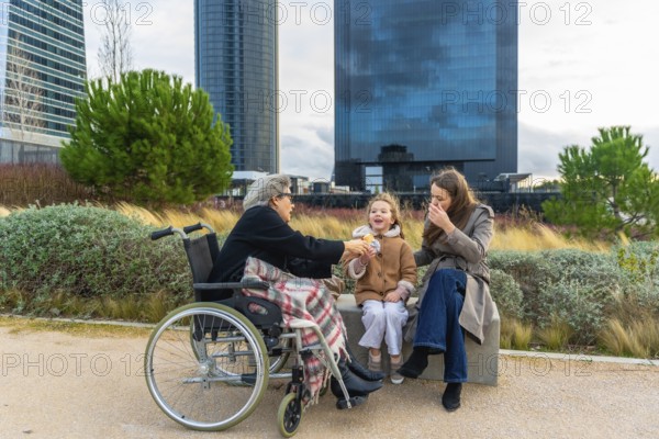 Grandmother in wheelchair enjoys a snack from her smiling granddaughter while family relaxes together outdoors in an urban park, sharing care, support and intergenerational bonding