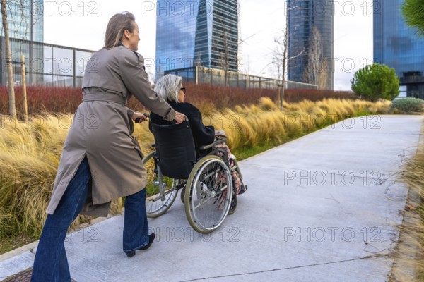 Caregiver pushing an elderly woman in a wheelchair along an outdoor path in a modern city park, providing support and companionship during a leisurely stroll