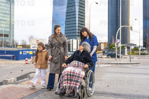 Family members and a professional caregiver are walking outdoors with a smiling senior woman in a wheelchair, enjoying a pleasant stroll in an urban environment surrounded by modern skyscrapers