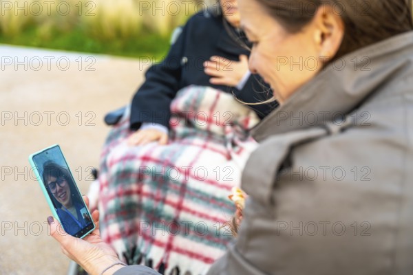 Daughter visiting grandmother in a wheelchair outdoors, sharing a video call with another family member, highlighting connections and communication across generations