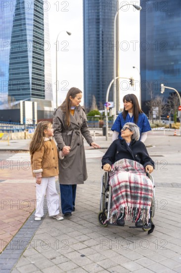 Family members and a caregiver are walking with an elderly woman in a wheelchair, providing care and companionship during an outdoor stroll in a modern city setting