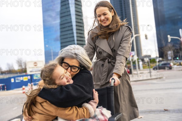 Granddaughter embraces her grandmother sitting in a wheelchair while a woman pushes them, enjoying an outdoor stroll in an urban setting with modern buildings