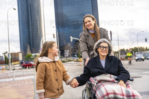 Three generations walking in the city, grandmother in a wheelchair, daughter pushing and granddaughter holding hands, sharing care, warmth and joyful outdoor togetherness