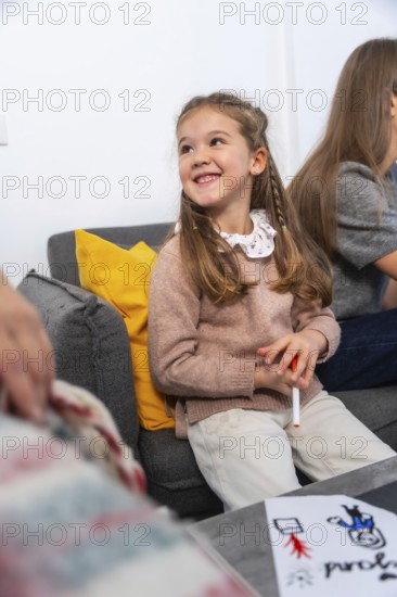 Happy little girl sitting on a sofa, smiling while holding a marker and showing a homemade drawing depicting a person in a wheelchair, sharing a moment with family