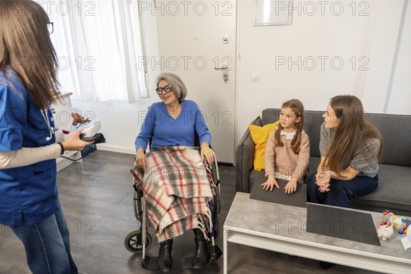Nurse communicating with an elderly woman in a wheelchair, while her daughter and granddaughter are visiting, focusing on elder care and family support in a home environment