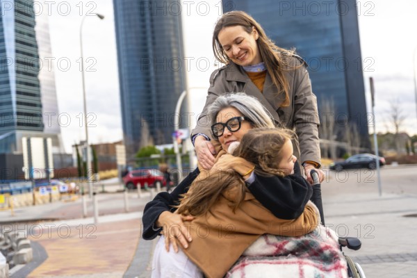 Multi generational family group spending time together outdoors, with a daughter pushing her mother in a wheelchair while the granddaughter hugs her, showing love, care, and support