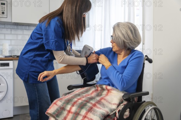 Healthcare professional in blue scrubs measuring an elderly woman's blood pressure in a wheelchair at home, providing attentive medical care and support for daily wellbeing