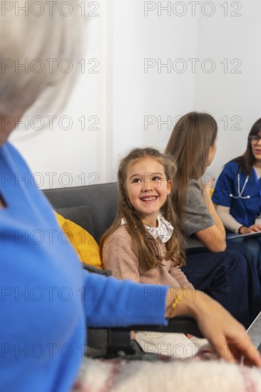 Smiling young girl visits grandmother in a wheelchair at a nursing home, surrounded by family and a caring healthcare professional sharing warmth, support and conversation