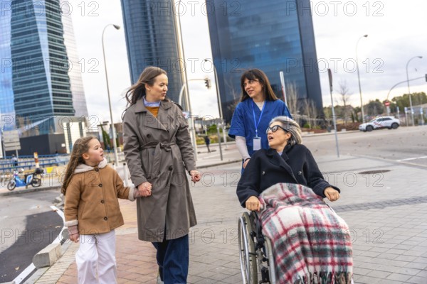 Family members and a caregiver pushing an elderly woman in a wheelchair, leaving a healthcare facility for a walk along a city street lined with modern buildings