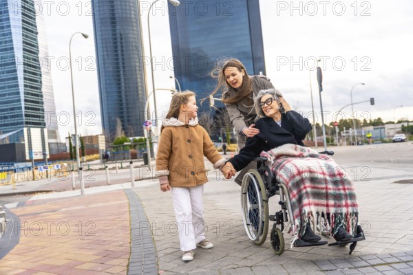 Multi generational women sharing a city walk, grandmother in wheelchair holding granddaughter's hand as mother pushes, smiling and connecting in winter coats under urban skyline