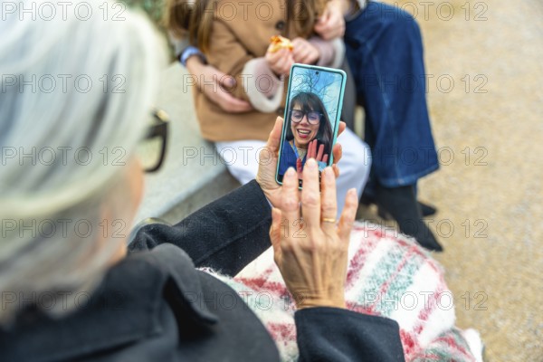Elderly woman in a wheelchair using a smartphone for a video call with a smiling relative, staying connected with family while enjoying an outdoor walk