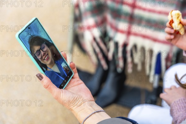 Hand holding a smartphone displaying an elderly woman on a video call, staying connected with a person in a wheelchair and a child's hand holding food in the background