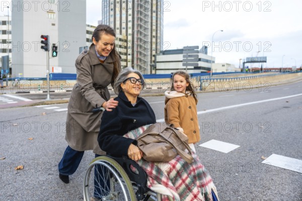 Family members assisting senior woman in wheelchair outdoors, granddaughter walking alongside while navigating city street, representing care, support, and active lifestyle for elderly