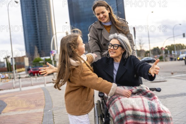 Smiling grandmother in wheelchair reaching to embrace granddaughter while adult woman pushes them along a sunny city sidewalk, showing intergenerational care, support and joy