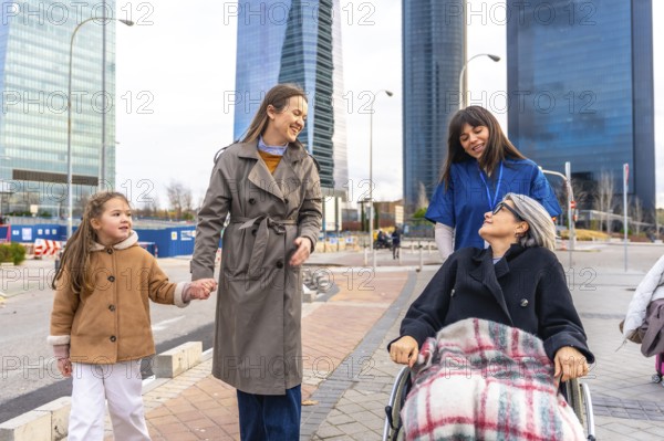 Caring caregiver pushing a senior woman in a wheelchair outdoors, joined by her daughter and granddaughter, sharing moments of companionship and happiness in an urban environment