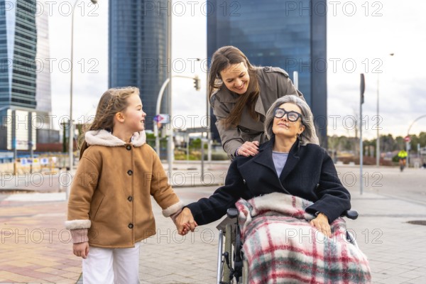 Three generations of women, including a senior woman in a wheelchair, a smiling adult daughter pushing her, and a young girl holding hands, sharing a happy moment during an outdoor stroll in a city