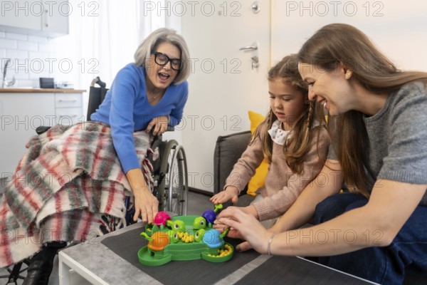 Senior woman in a wheelchair. Her daughter. And granddaughter happily playing a hungry hungry hippos style board game together. Celebrating connection. Family bonding. And inclusive leisure activities