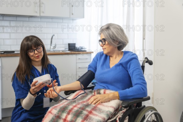 Nurse using a digital device to check blood pressure on arm of an elderly woman seated in a wheelchair, providing healthcare support and monitoring vital signs during a home visit