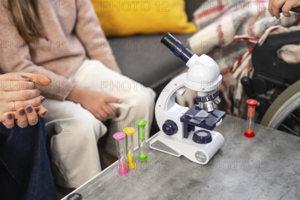 Granddaughter and family explore science together with a microscope and sand timers during a warm visit with grandmother in a wheelchair, sharing learning, care, and connection