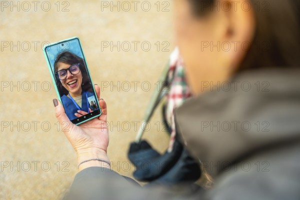 Person holding smartphone showing video call with smiling woman while pushing a wheelchair outdoors, caregiver connecting family and elderly through technology for support and companionship