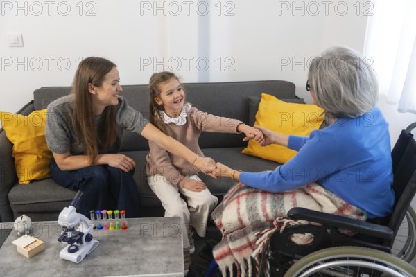 Three generations of women, a cheerful child, her mother, and a senior grandmother in a wheelchair, are connecting and holding hands, expressing care, support, and strong family bonds during a visit