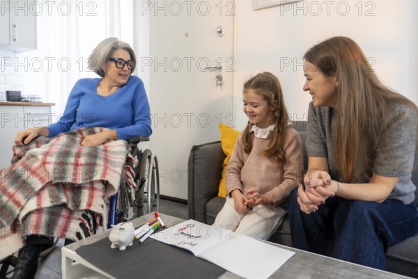 Family members showing love and care visiting an elderly woman sitting in a wheelchair while granddaughter draws at home, sharing happy moments together