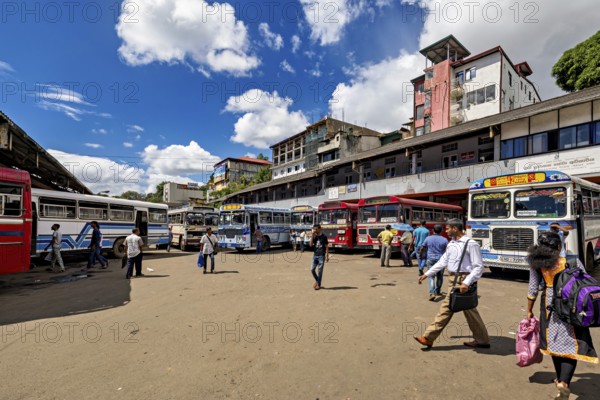 People walk past a bus stop surrounded by various buses and clouds in the sky, Kandy bus station in Sri Lanka
