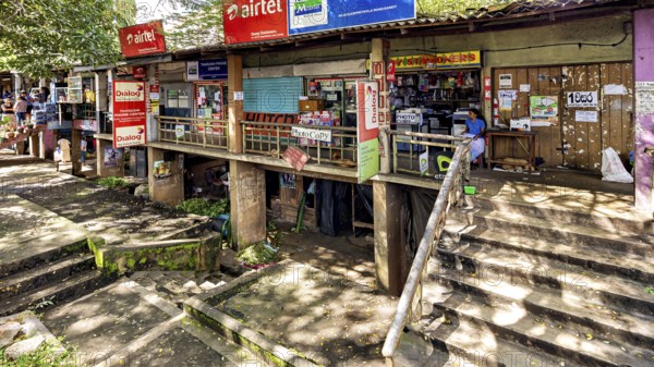 A storefront with various signs and posters surrounded by trees and a rustic atmosphere, The old market halls of Kandy in Sri Lanka