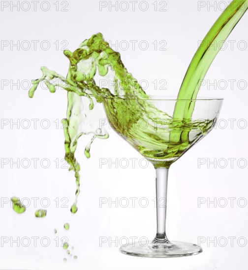 A powerful flood of green liquid pours into a cocktail glass and spills over, studio shot against a white background