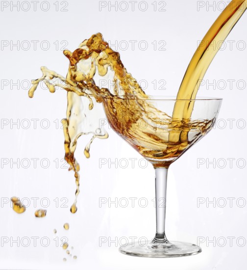 A strong flood of golden yellow liquid pours into a cocktail glass and spills over, studio shot against a white background