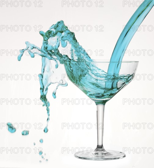 A powerful flood of blue liquid pours into a cocktail glass and spills over, studio shot against a white background