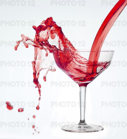 A powerful flood of red liquid pours into a cocktail glass and spills over, studio shot against a white background