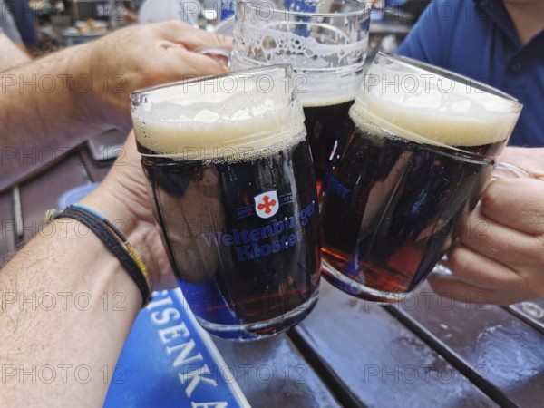 Three people toast in a beer garden with dark beer glasses, cheerful atmosphere, Weltenburg Abbey, Bavaria