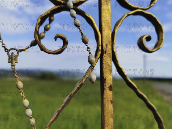 Close-up of a metal religious ornament, part of a cross, with a meadow and sky in the background, Frankenwald nature park Park