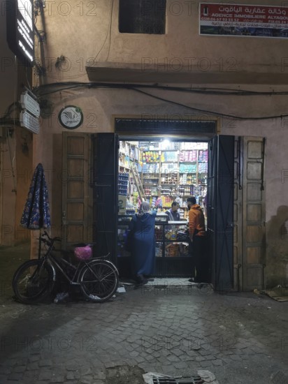 Small shop at night with open gate and bicycle, passers-by on the street in the Medina, Marrakech, Morocco