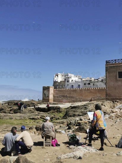 Painters paint the city view on the coast in front of old city walls and rocks under a blue sky, Essaouira, Marakesh