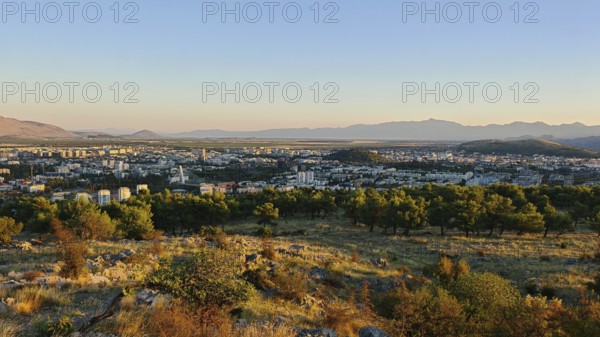 A city surrounded by nature with trees in the foreground and mountains in the background, illuminated by a warm sunset, looking out from a hill on the outskirts of Podgorica, Montenegro