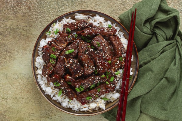 Crispy beef, with green onions and sesame seeds, on a rice pad, in a bowl with chopsticks, on a textured surface, no people