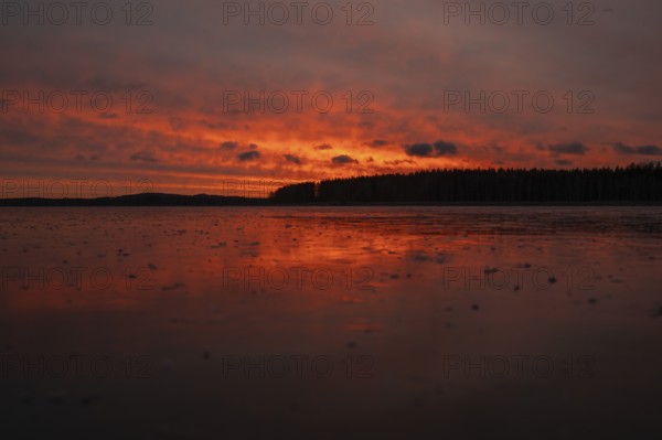 A dramatic sunset over a still, frozen lake and forest with orange-red hues in a cloudy sky, Finland