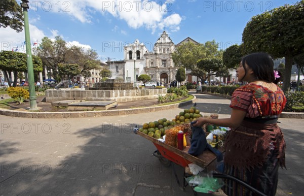 Parque Centroamérica or Parque Central, a Mayan woman in traditional dress selling oranges, in the back the cathedral or Catedral del Espíritu Santo, Quetzaltenango or Xela, Quetzaltenango province, Guatemala
