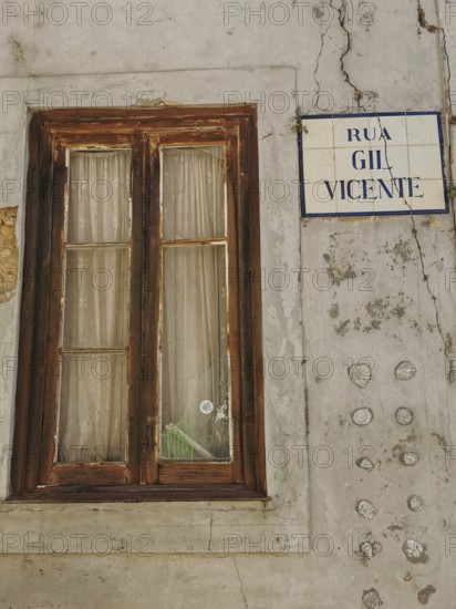 Old window with wooden frame and curtains next to a street sign on a rough, weathered wall