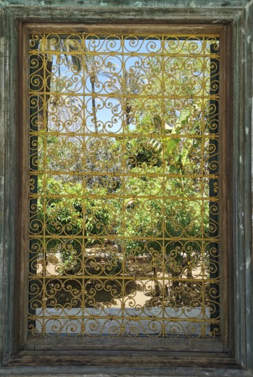 A richly decorated iron window provides a view of a green garden under a clear sky