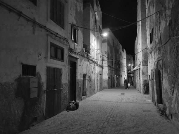 Nighttime scene of a quiet alley illuminated by a few alleyway lamps in black and white, Essaouira, Morocco
