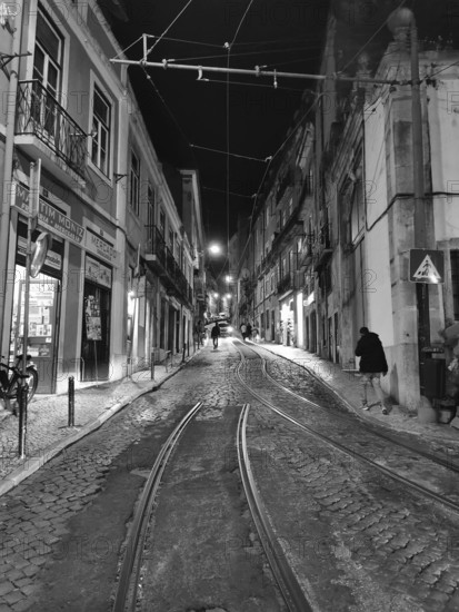 Night view of a paved street with tram tracks and illuminated buildings in black and white, Lisbon, Portugal