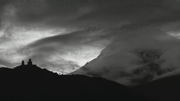 Dramatic silhouette of church buildings in front of a cloud-covered mountain, Kazbegi, Stepantsminda, Georgia