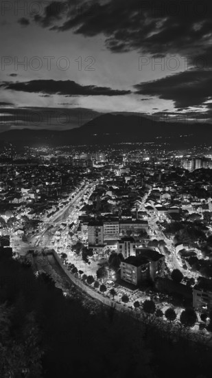 Night view of an illuminated city with mountains in the background under a cloudy sky in black and white, Prizren, Kosovo