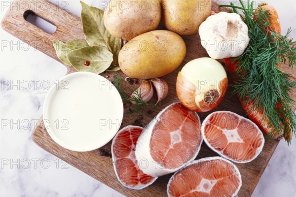 Potatoes, salmon, garlic, onion, and herbs are placed on a wooden cutting board. These ingredients are ready for meal preparation in a kitchen