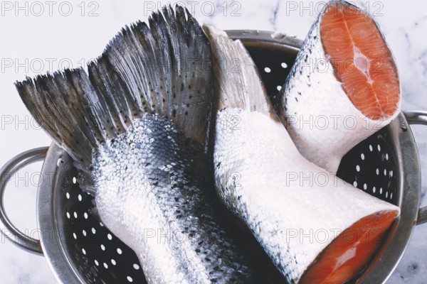 Fish tails are placed in a colander on a marble counter. The colander allows water to drain away from the fish. The fish looks fresh and clean, ready for cooking or preparation