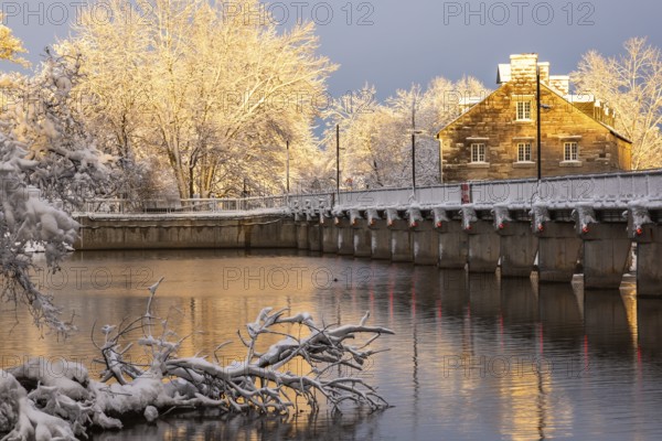 Moulin Neuf water flow control dam and walkway over Des Mille-Iles river plus New Mill on Ile des Moulins with first winter like snow in late autumn at sunset, Old Terrebonne, Quebec, Canada