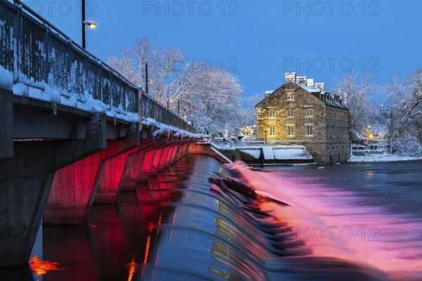 Illuminated Moulin Neuf water flow control dam and walkway over Des Mille-Iles river plus New Mill on Ile des Moulins with first winter like snow in late autumn at dusk, Old Terrebonne, Quebec, Canada